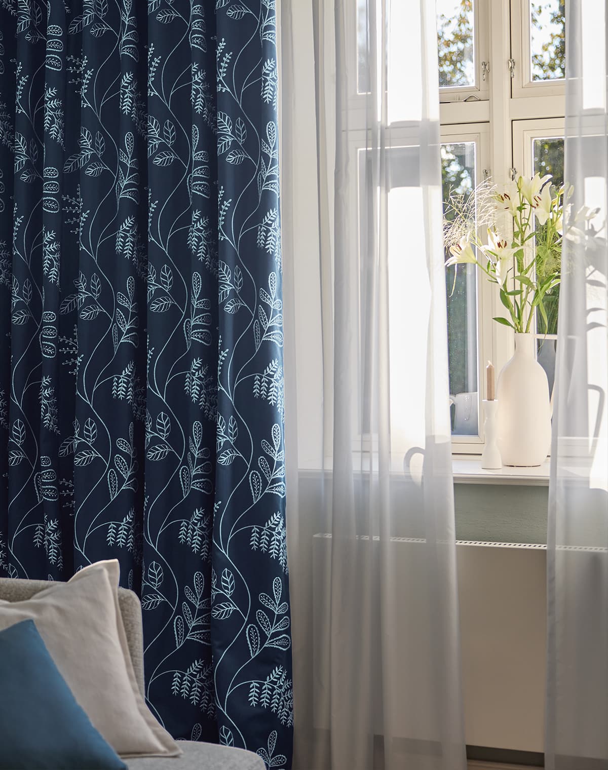 Eine blaue Gardine mit floralem Muster und eine weiße Gardine am Fenster.