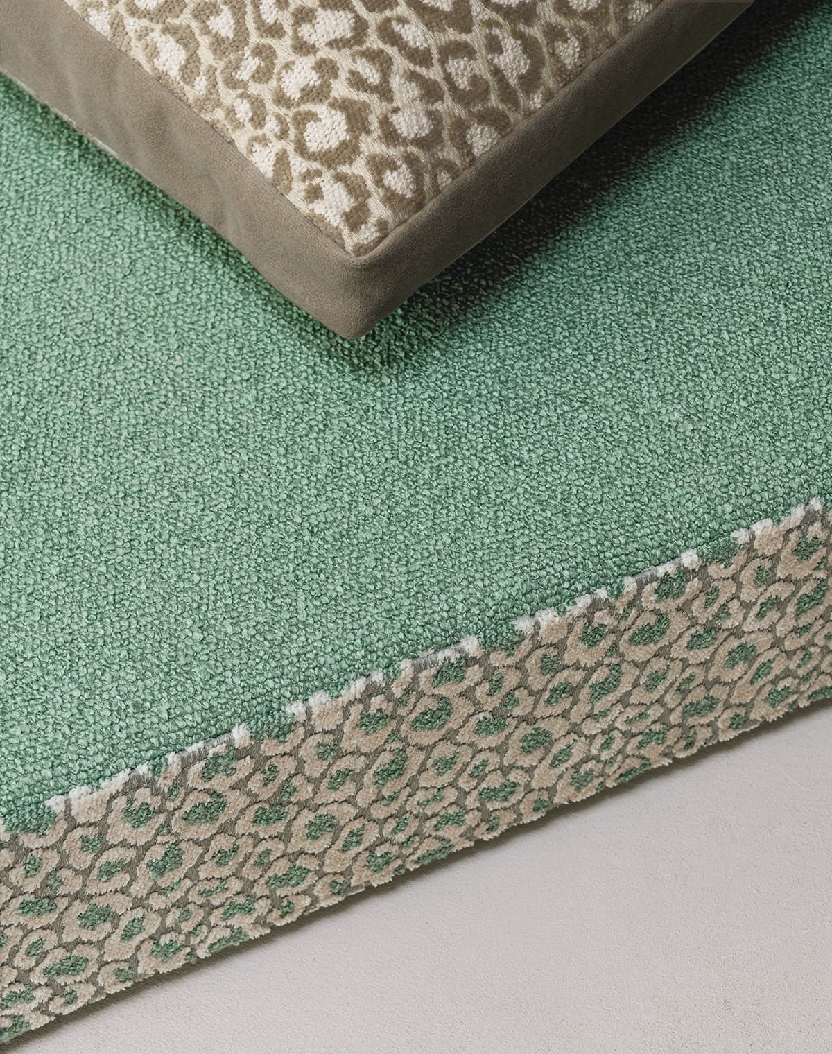Das Bild zeigt Detailaufnahme eines Grünen Polsters. Das Polster hat eine mehr weiß grüne Farbe. Mit einer Bemusterung an der Seite. Auf dem Polster liegt ein Kissen in braun weiß, dass an das Muster des Polster erinnert.