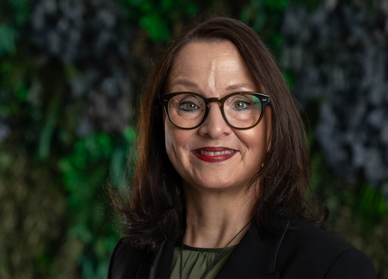 Portraitfoto von einer Frau mit braunen Haaren mit Brille. Sie steht vor einer grünen Wand und lächtelt in die Kamera.