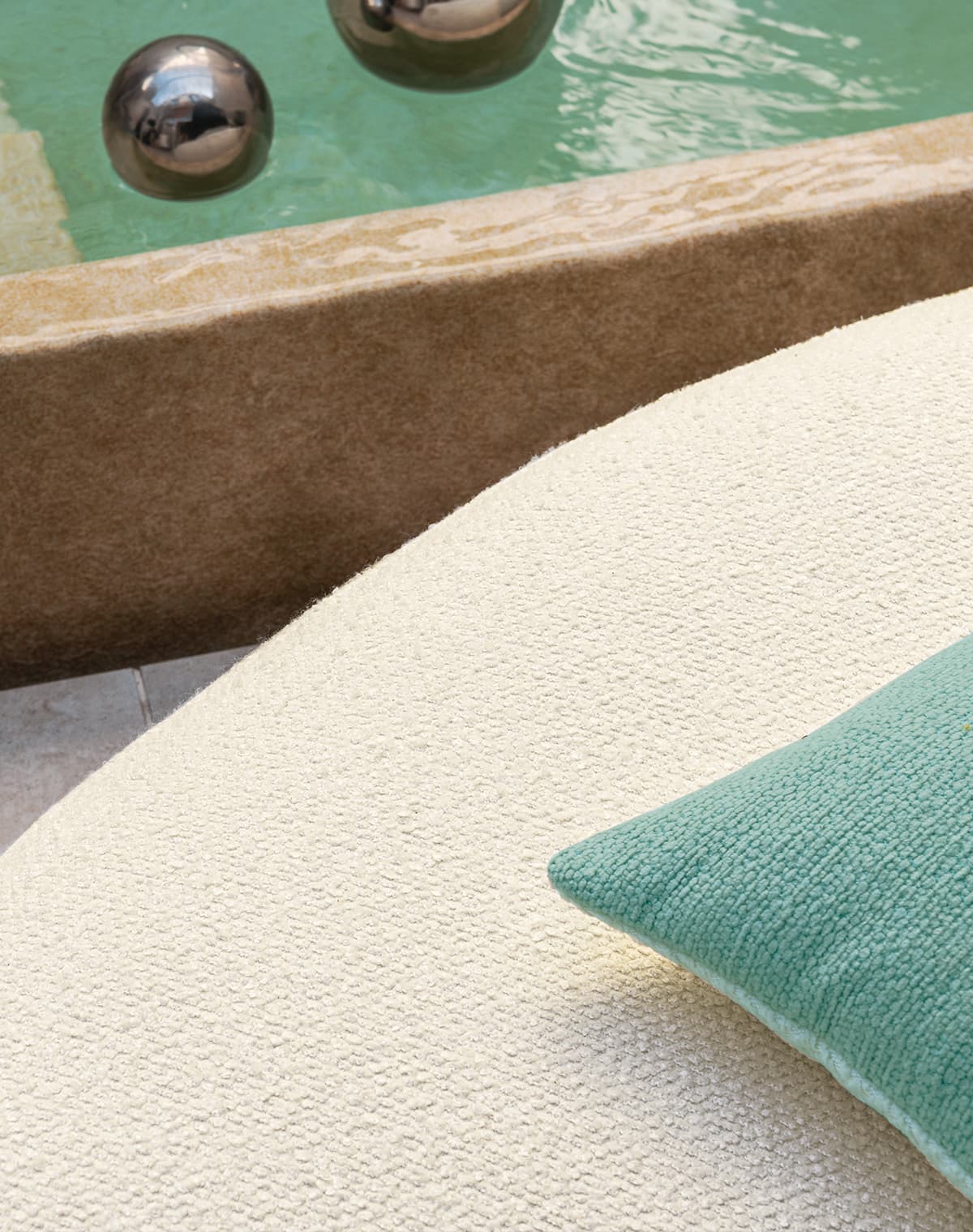 Das Bild zeigt eine Detailaufnahme von Outdoor-Stoffen, mit einem Ausschnitt eines gelb-weißen Sofas und einem türkisen Kissen. Im oberen Rand ist ein Teil eines Naturpools zu erkennen, dessen grün-türkise Farbe wunderbar zu dem Kissen passt.