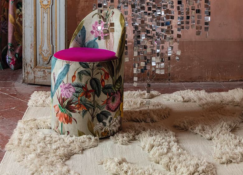 Das Bild zeigt einen Pouf-Hocker mit floralem Design auf einem weißen Teppich. Der Teppich hat eine flauschige Struktur und ein klassisches Gewebe. Die Umgebung wirkt kühl, wird jedoch durch den Hocker und den Teppich wieder warm und einladend.
