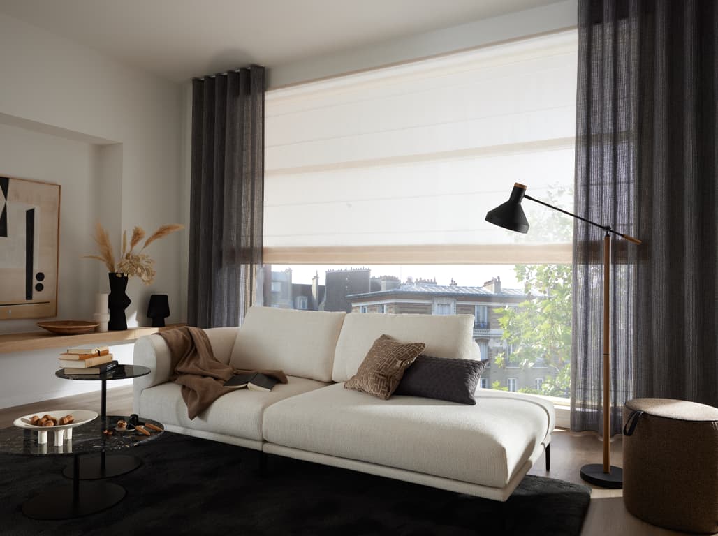 Faltrollo Summer Season in creme in einer Raumsituation mit einem weißen Sofa vor dem Fenster, einer Stehlampe, einem Hocker, einem  schwarzen Teppich und grauen Vorhängen