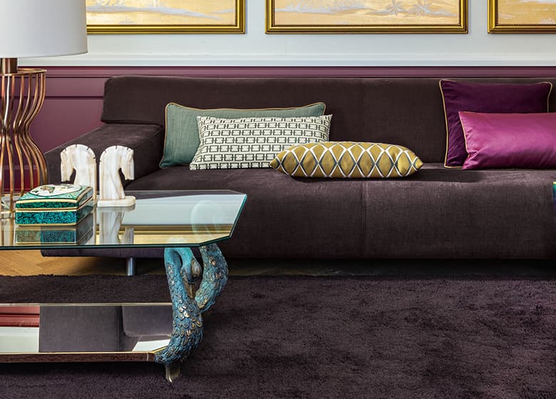 Das Bild zeigt einen lila Teppich und ein Sofa im Wohnzimmer, dekoriert mit einem Tisch und vielen Kissen. Die Kombination aus Teppich und verschiedenen Stoffen schafft eine gemütliche Atmosphäre.