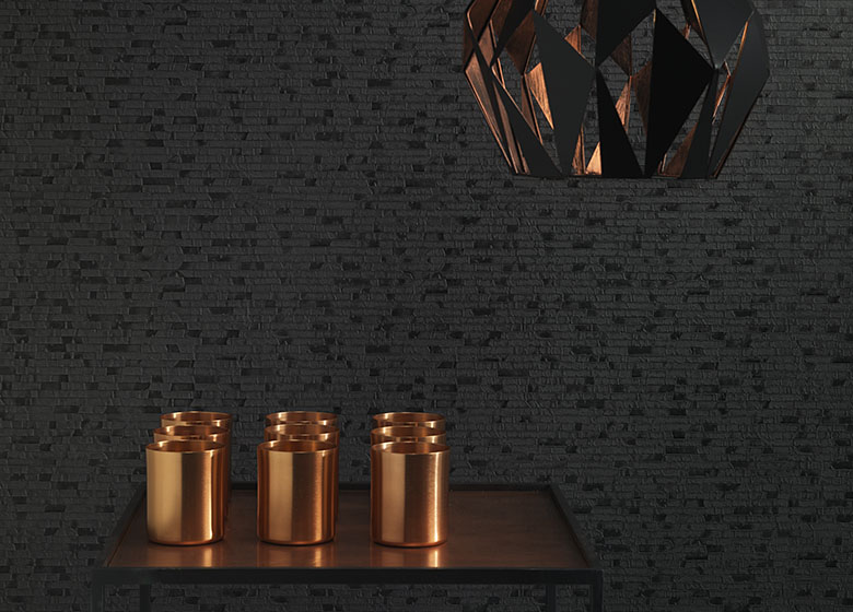 Das Bild zeigt ein dunkles Wanddesign mit dekorativen Artikeln in kupferfarbenem Glanz im Vordergrund. Ein passender Lampenschirm in Kupferglanz von der Decke setzt einen spannenden Kontrast.