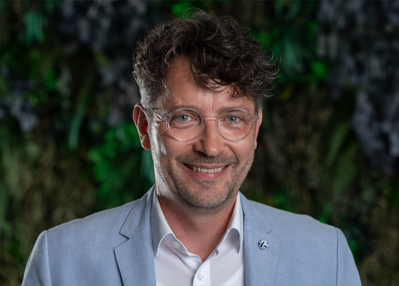 Portraitfofo von einem Mann mit braunen Haaren und runder Brille vor einer Pflanzenwand. Er lächelt freundlich in die Kamera.