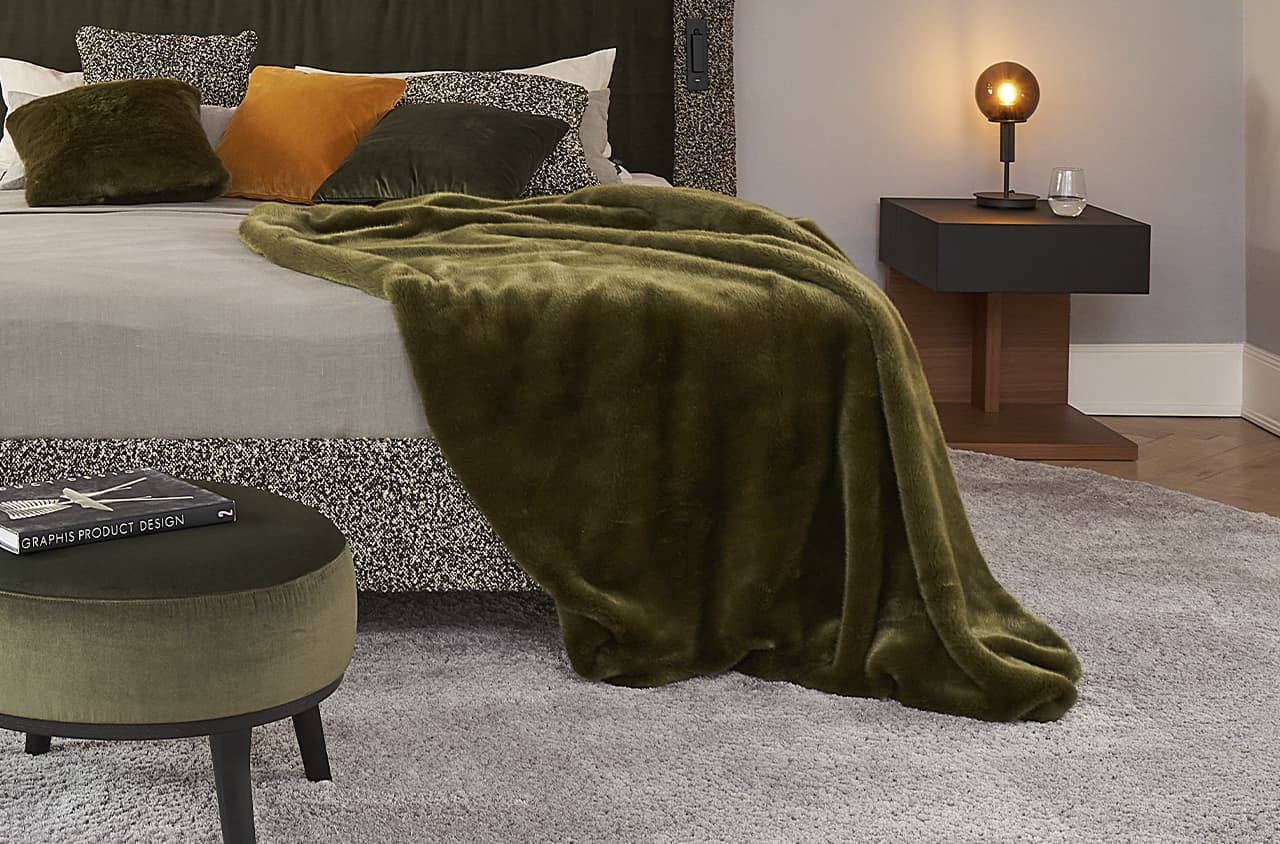 Eine grüne Kuscheldecke liegt af einem grauen Polsterbett und hängt auf den Boden herunter.