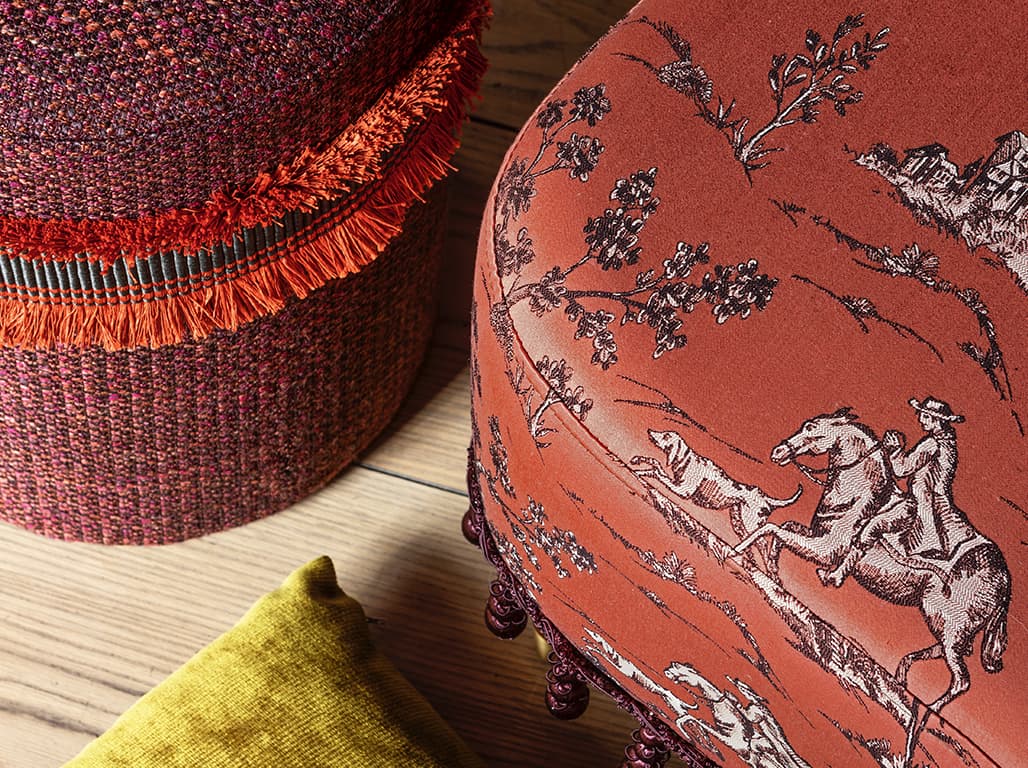 Das Bild zeigt eine Detailaufnahme eines Poufs in Lila und eines orangefarbenen Hockers mit Barockmustern. Die verschiedenen dunkellila und dunkelorange Töne harmonieren auf besondere Weise miteinander.