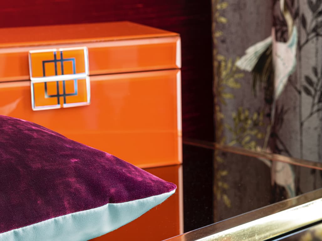 Das Bild zeigt eine Detailaufnahme eines lila und hellblauen Kissens. Im Hintergrund steht eine kräftig orangefarbene Box, die einen starken Kontrast zu den sanften Farben des Kissens bildet.