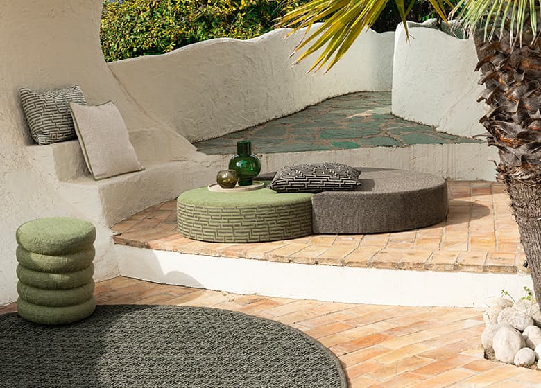 Das Bild zeigt einen Teppich auf einer Terrasse, umgeben von zahlreichen Dekorationsartikeln in verschiedenen Farben und Formen. Die geschwungene Architektur der Terrasse schafft zusammen mit dem Teppich eine einladende Atmosphäre.