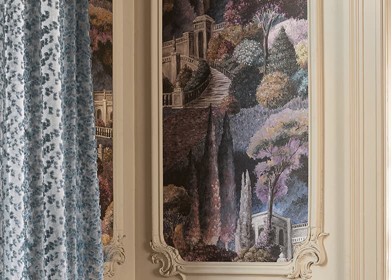 Das Bild zeigt eine blaue Gardine und eine Tapete mit Printmotiv. Das Blau der Gardine spiegelt sich dezent in der Tapete wider.